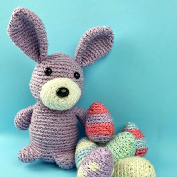 Lavender crocheted bunny with a pile of colorful crocheted Easter eggs made with a free pattern from Shiny Happy World.