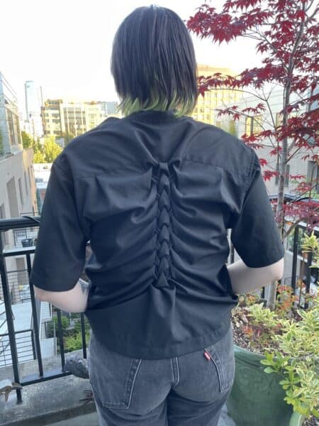 Back view of a person wearing a black smocked shirt on a balcony with cityscape background.
