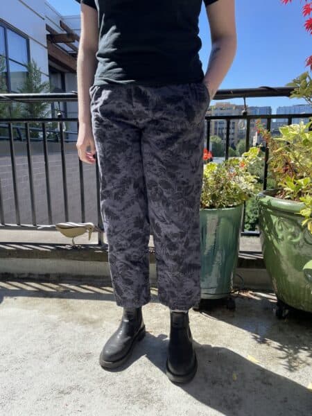 Person wearing black top, patterned pants, and black boots on balcony with plants.